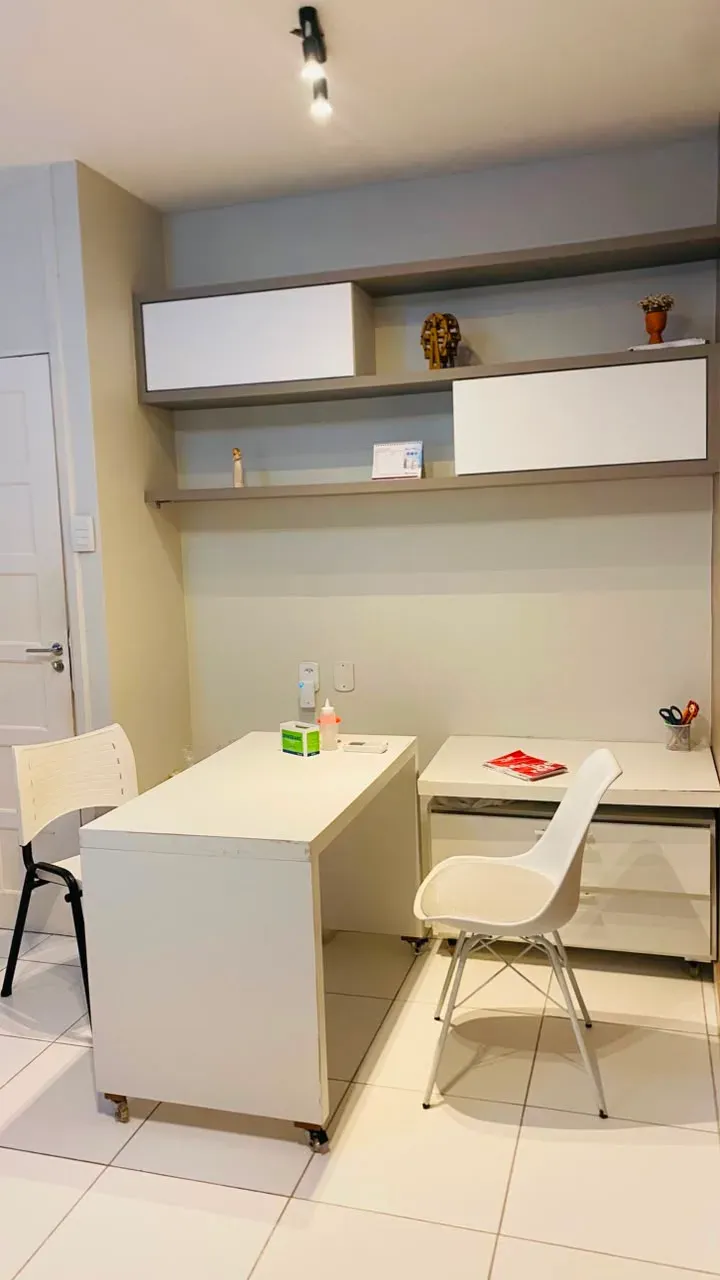 Consultório com mesa branca, cadeiras e prateleiras.