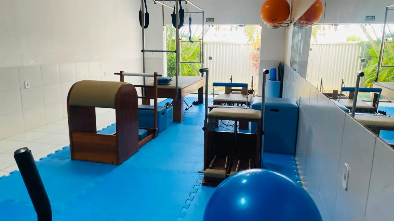 Outro ângulo do estúdio de pilates, com barrel, cadeira, bola azul e janela ao fundo.